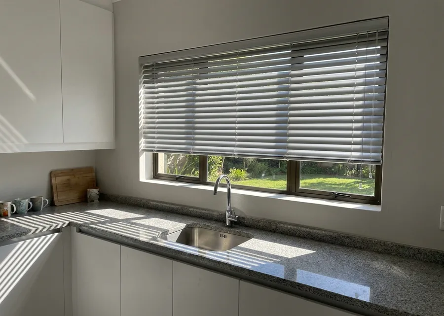 Silver aluminium venetian blind tilted open above the kitchen sink of a Menlo Park home, morning light striping the stone counter