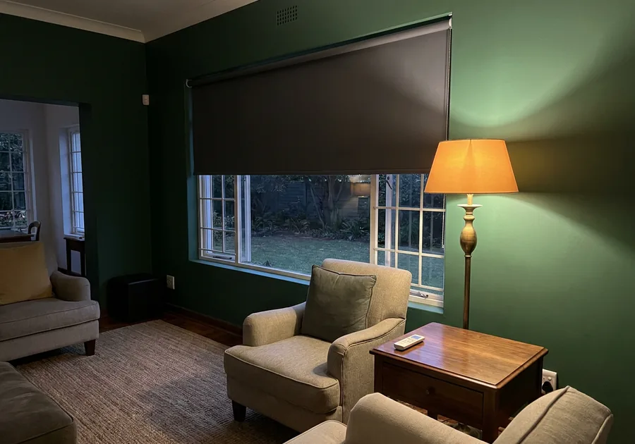 Motorised blockout roller part-lowered in a green-walled Menlo Park sitting room at lamplight, remote handset on the table
